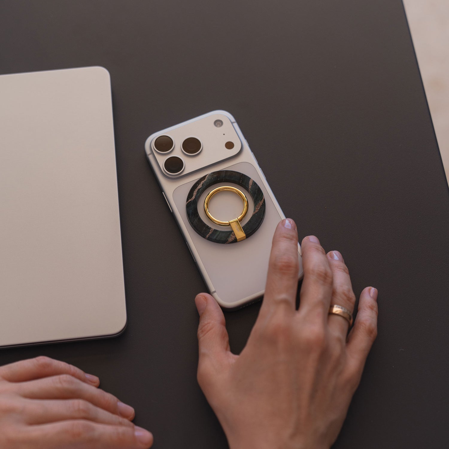 Eine Hand mit einem Goldring ruht neben einem arktis Magnetischer Ringhalter für MagSafe - Sweet Leopard, abgebildet mit iPhone Zubehör und einem geschlossenen Laptop auf einer dunklen Fläche.