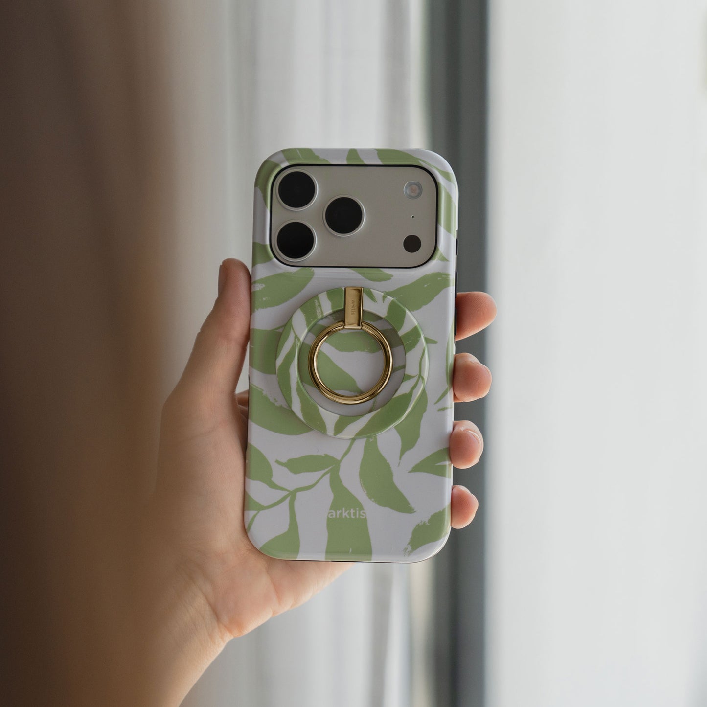 Eine Hand hält ein iPhone mit dem arktis Magnetischer Ringhalter für MagSafe - Botanic: eine stylische Hülle mit grünem Blattmuster und goldenem Ring mit starken N52 Magneten, die Stil und Funktion für Top-iPhone-Zubehör verbinden.