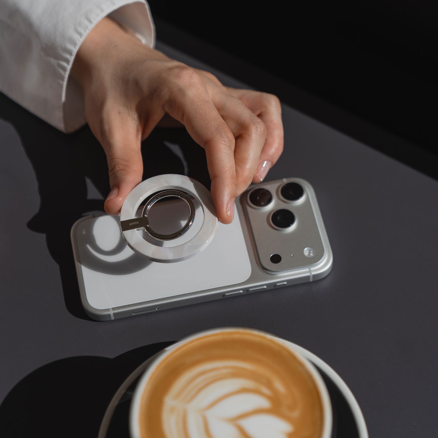 Eine Hand befestigt den arktis Magnetischer Ringhalter für MagSafe - Sweet Leopard mit starken N52 Magneten an einem iPhone auf einem Tisch neben einer Latte-stylisch und praktisch als angesagtes iPhone Zubehör.