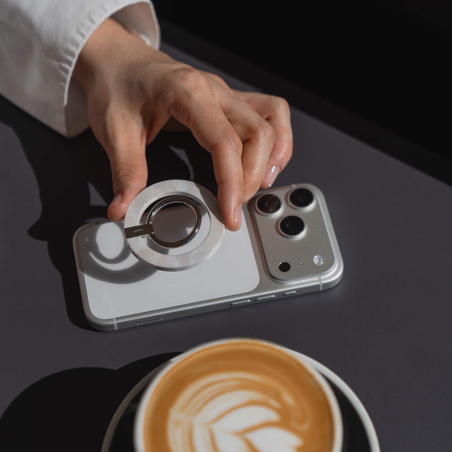 Eine Hand befestigt den arktis Magnetischer Ringhalter für MagSafe - Sweet Leopard mit starken N52 Magneten an einem iPhone auf einem Tisch neben einer Latte-stylisch und praktisch als angesagtes iPhone Zubehör.