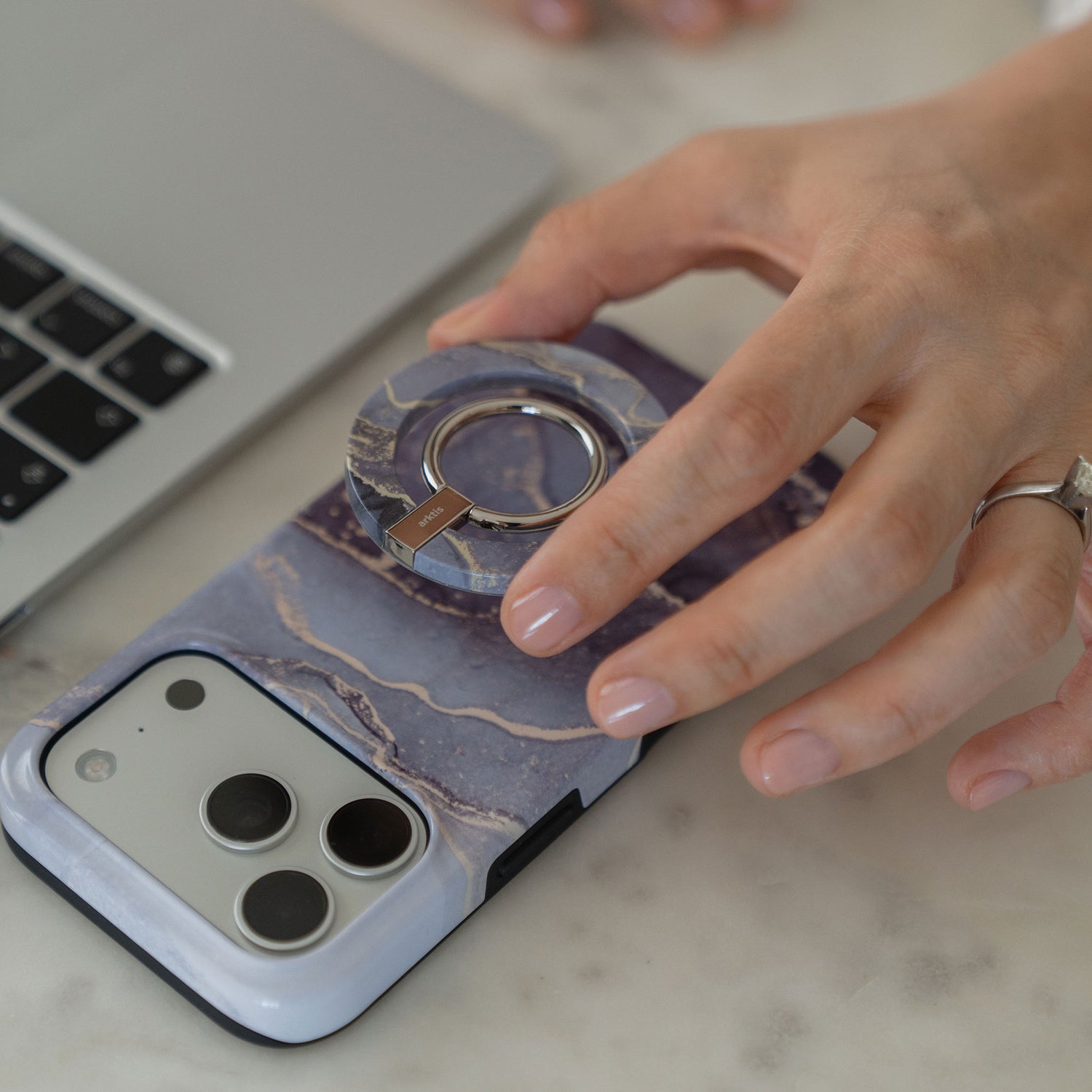 Eine Hand mit lackierten Fingernägeln hält die arktis iPhone 14 Pro Max Hülle - Dusty Marble, eine MagSafe-kompatible, stoßabsorbierende TPU-Silikonhülle mit einem Ringständer mit Marmormuster, neben einem Laptop auf einem Tisch.