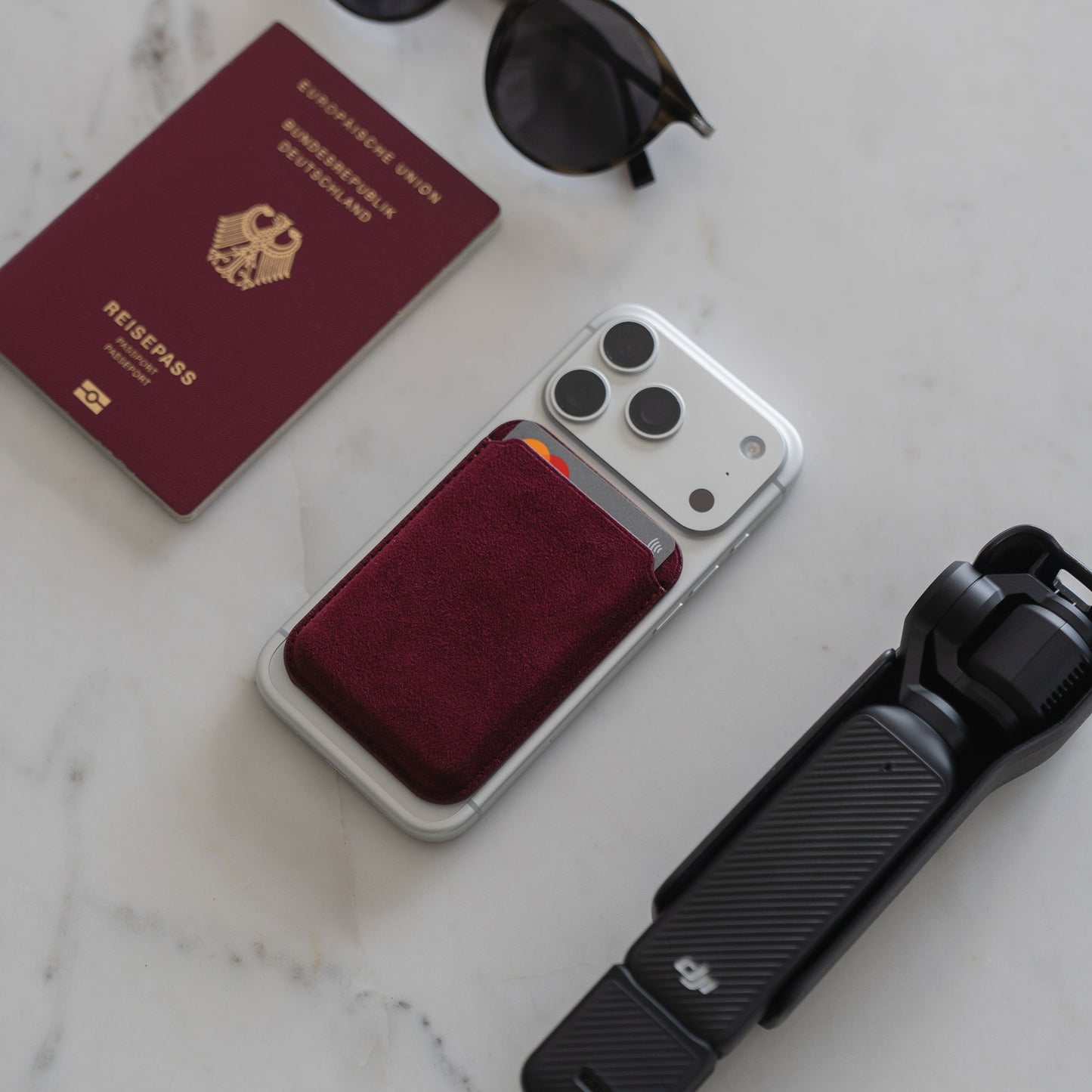 Auf einer weißen Fläche liegen ein deutscher Reisepass, eine Sonnenbrille, eine Kamera und ein iPhone mit dem arktis iPhone MagSafe Kartenhalter aus Alcantara von arktis.