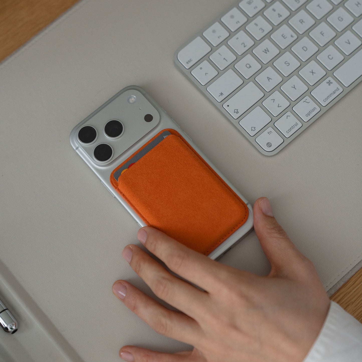 Eine Hand greift nach einem Smartphone neben der weißen Tastatur, neben dem arktis iPhone MagSafe Kartenhalter aus Alcantara von arktis.