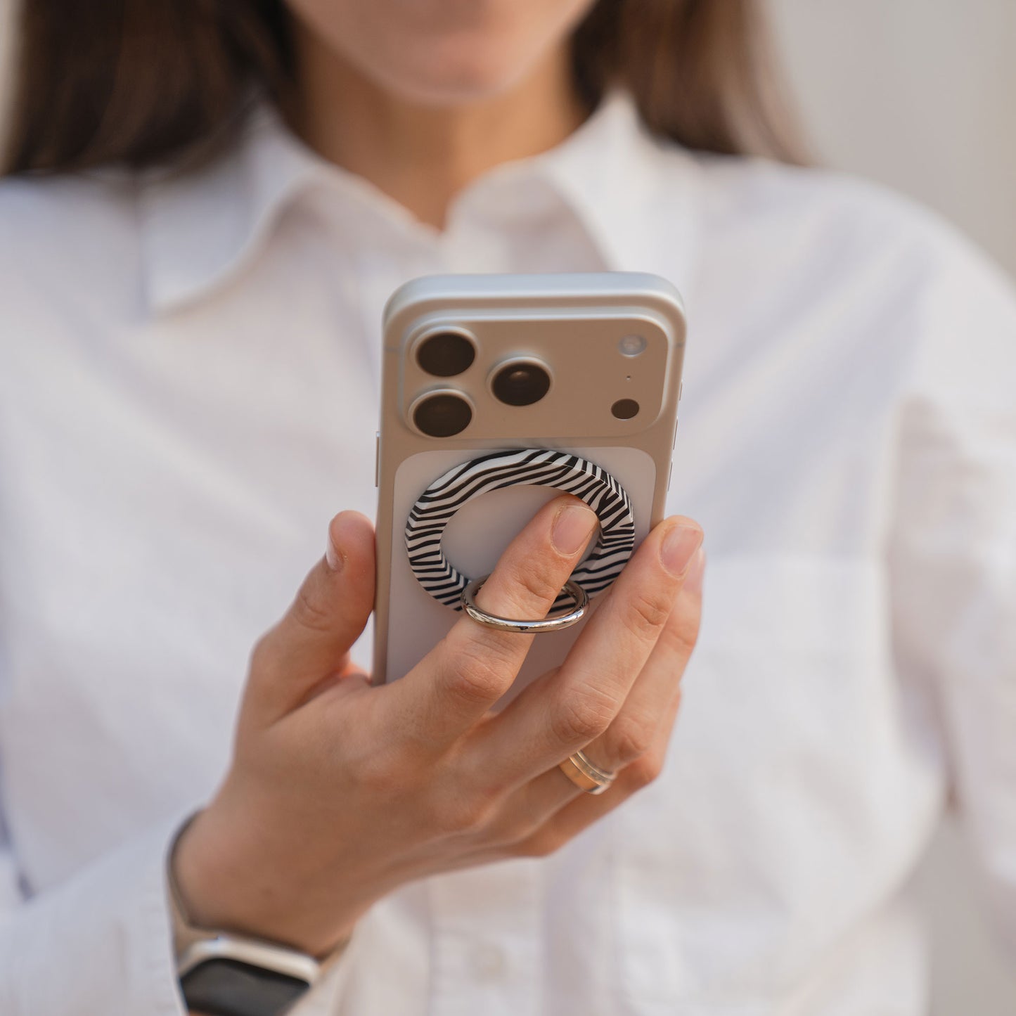 Eine Frau im weißen Hemd hält ihr Smartphone mit dem arktis Magnetischer Ringhalter für MagSafe - Sweet Leopard, mit starkem N52 Magneten - ein unverzichtbares iPhone Zubehör für Stil und Komfort.