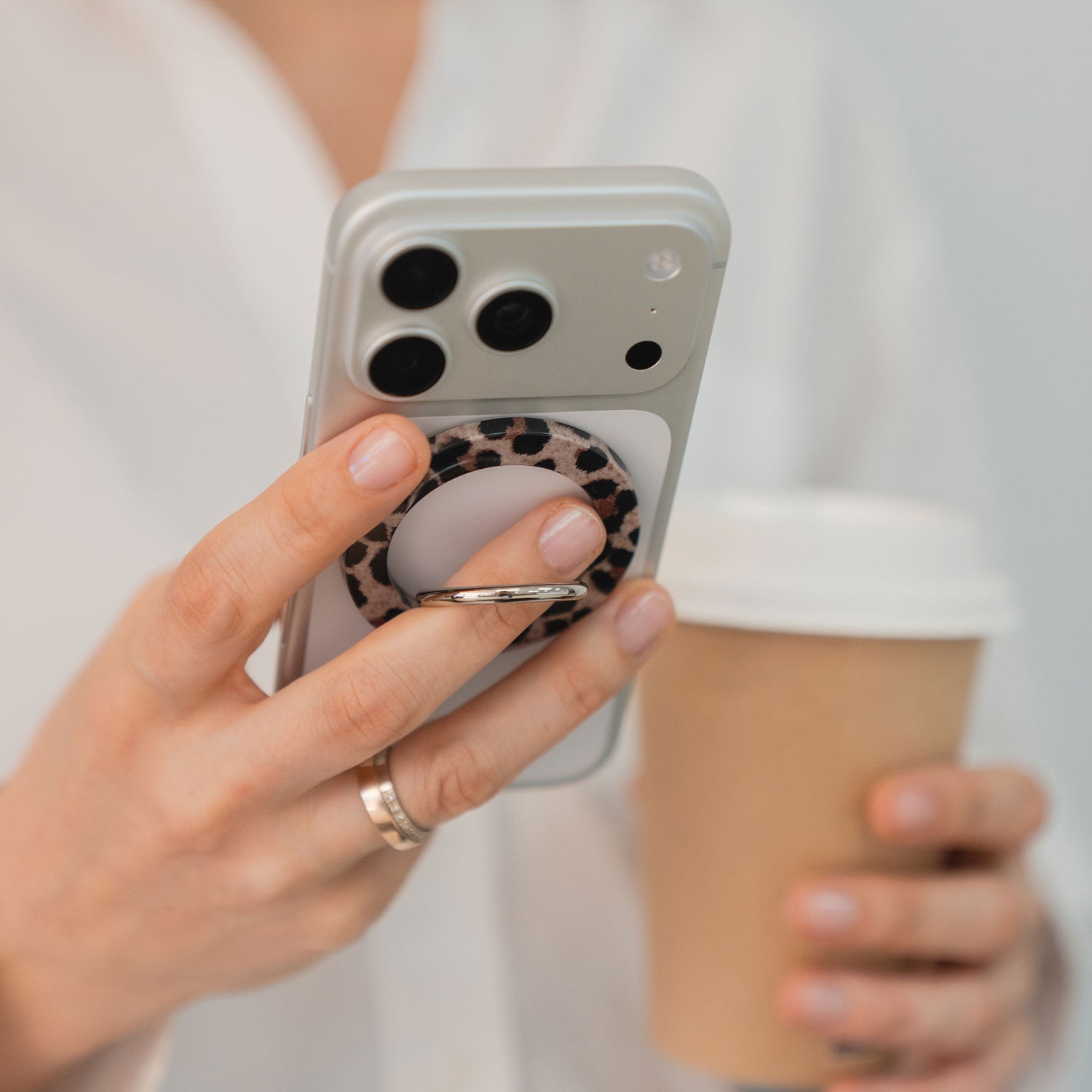 Eine Person hält einen arktis Magnetischer Ringhalter für MagSafe - Sweet Leopard als iPhone Zubehör, der an einem Smartphone befestigt ist, mit einem Kaffeebecher aus Papier und trägt ein weißes Hemd.