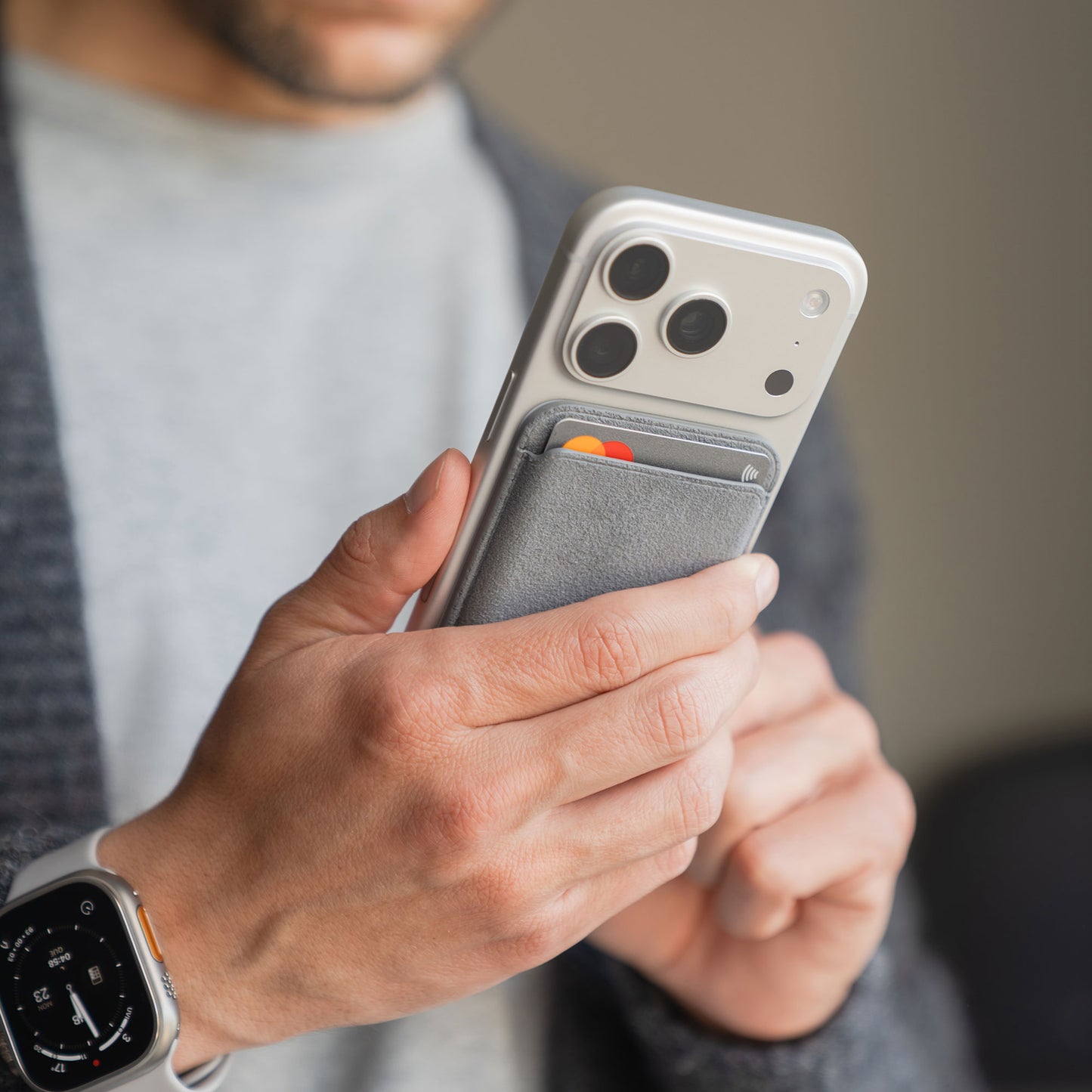 Eine Person hält ein iPhone mit dem arktis iPhone MagSafe Kartenhalter aus Alcantara in Grau, mit Karten im Inneren. Sie trägt außerdem eine Smartwatch und einen grauen Pullover.