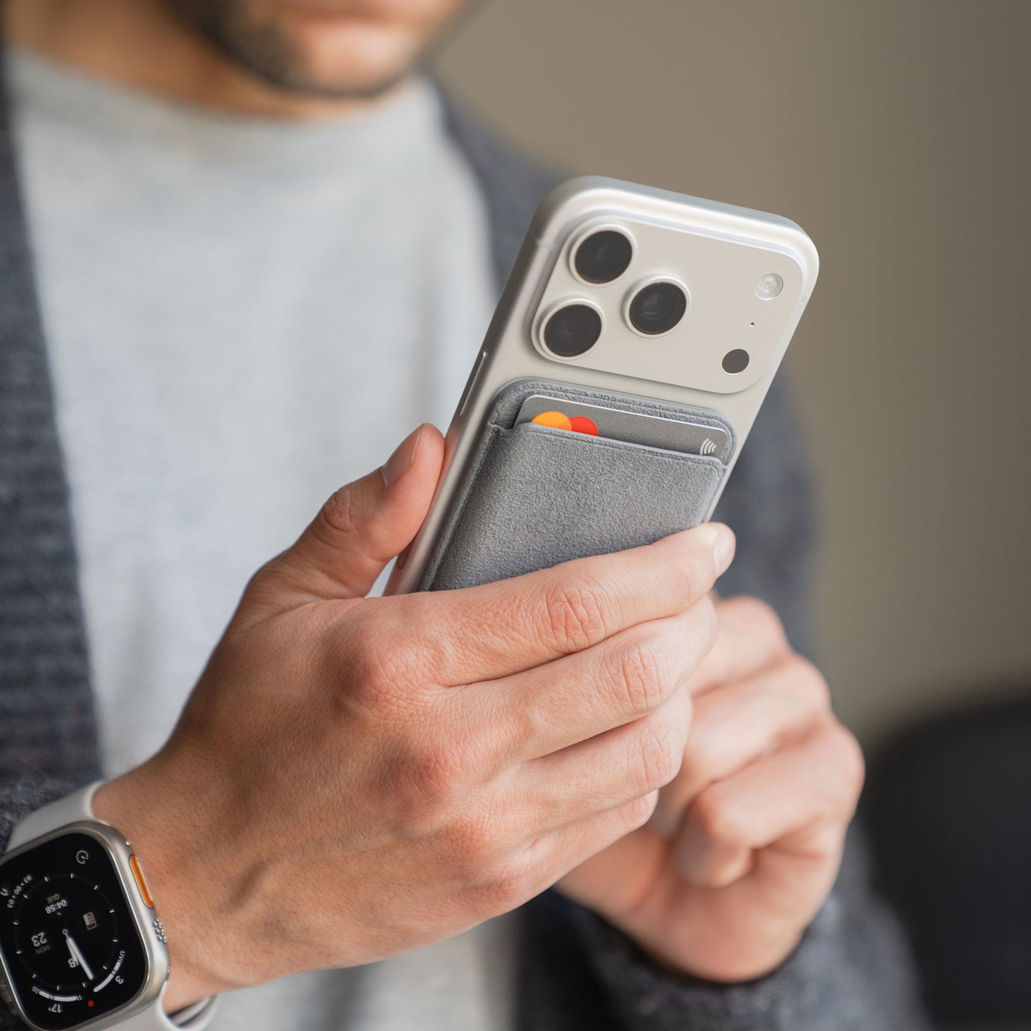 Eine Person hält ein iPhone mit dem arktis iPhone MagSafe Kartenhalter aus Alcantara in Grau, mit Karten im Inneren. Sie trägt außerdem eine Smartwatch und einen grauen Pullover.