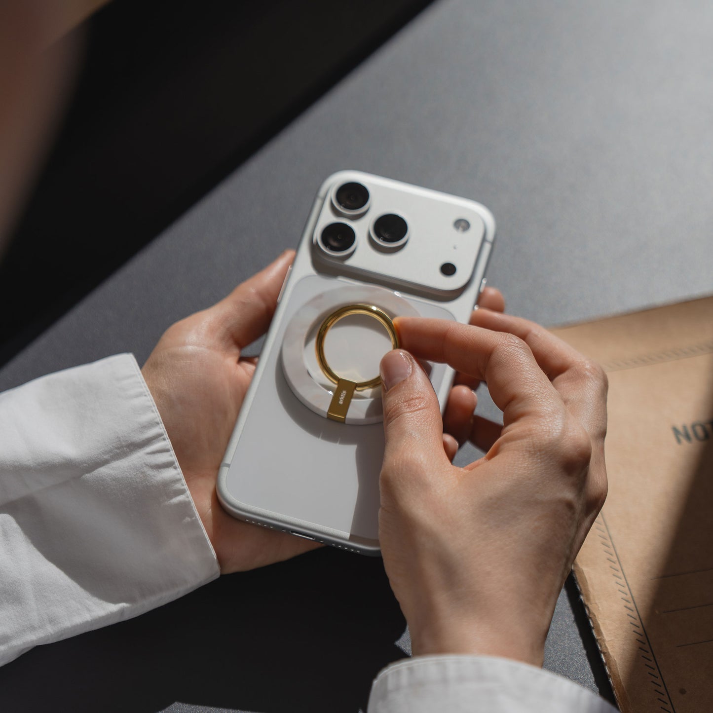 Eine Person befestigt den arktis Magnetischer Ringhalter für MagSafe - Sweet Leopard an einem weißen Smartphone auf einem schwarzen Tisch und präsentiert dabei stilvolles iPhone Zubehör mit starken N52 Magneten für einen sicheren Halt.