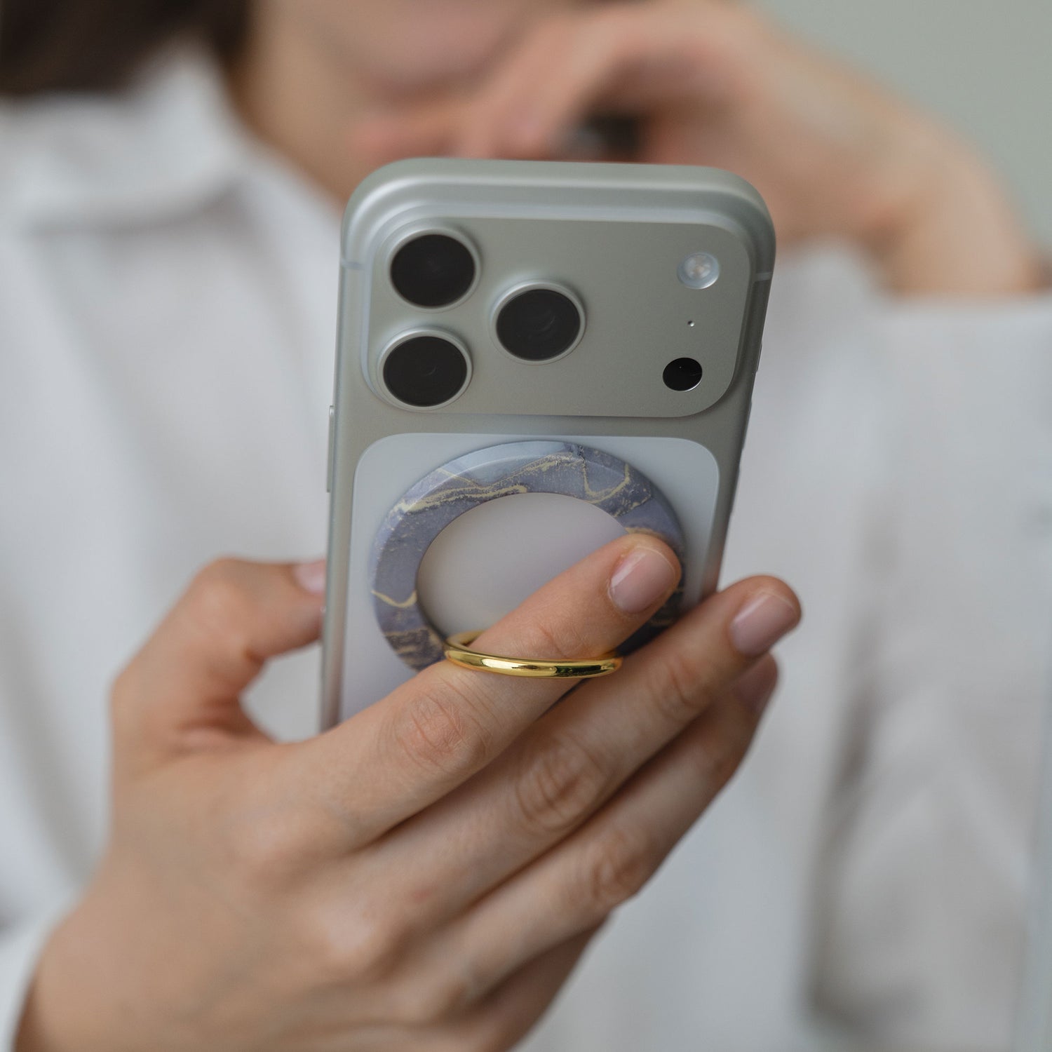 Eine Person hält ein iPhone mit dem arktis Magnetischer Ringhalter für MagSafe - Sweet Leopard, der mit starken N52-Magneten ausgestattet ist, und trägt einen goldenen Ring - eine stilvolle und funktionelle Zubehörwahl.