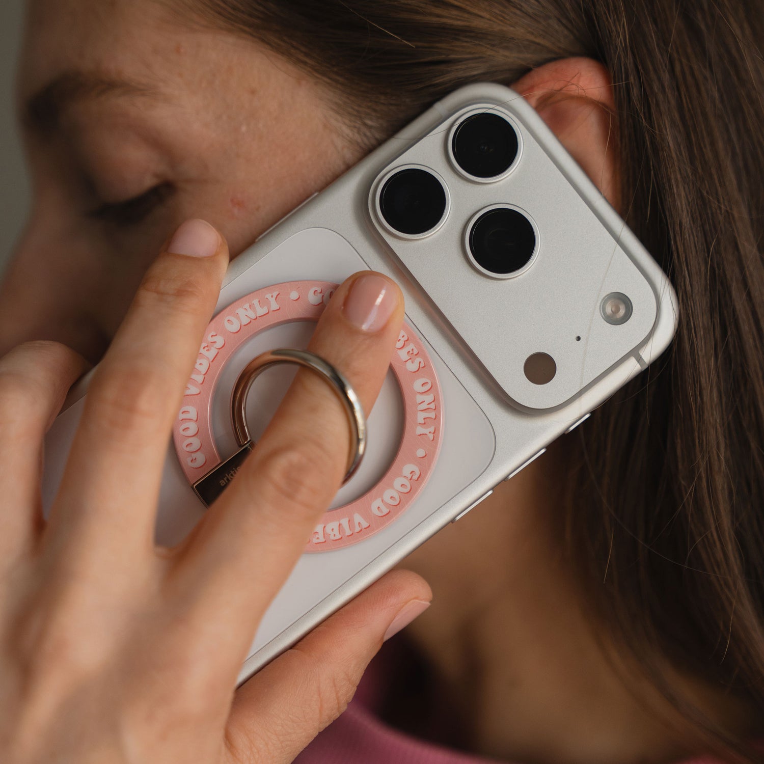 Eine Frau hält ihr Telefon mit dem arktis Magnetischer Ringhalter für MagSafe - Good Vibes an ihr Ohr, dem idealen Zubehör für sicheren Halt und positiven Style.
