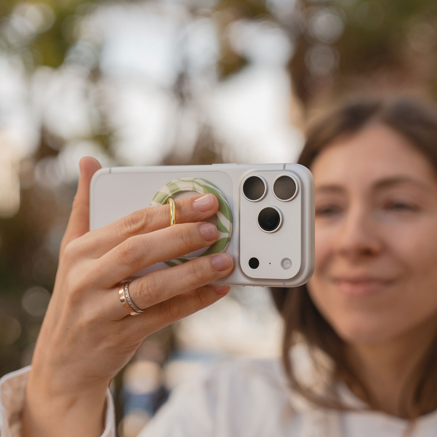 Eine Frau lächelt, während sie ein Selfie im Freien zwischen Bäumen macht. Sie hält ihr Smartphone mit dem arktis Magnetischer Ringhalter für MagSafe - Botanic in der Hand.
