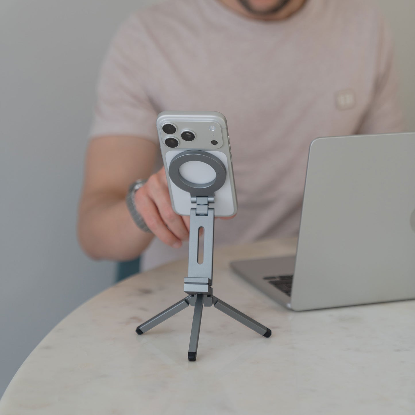 Eine Person benutzt ein Smartphone auf einem arktis iPhone MagSafe Tripod 360 Halterung von arktis neben einem Laptop auf einem runden Marmortisch.
