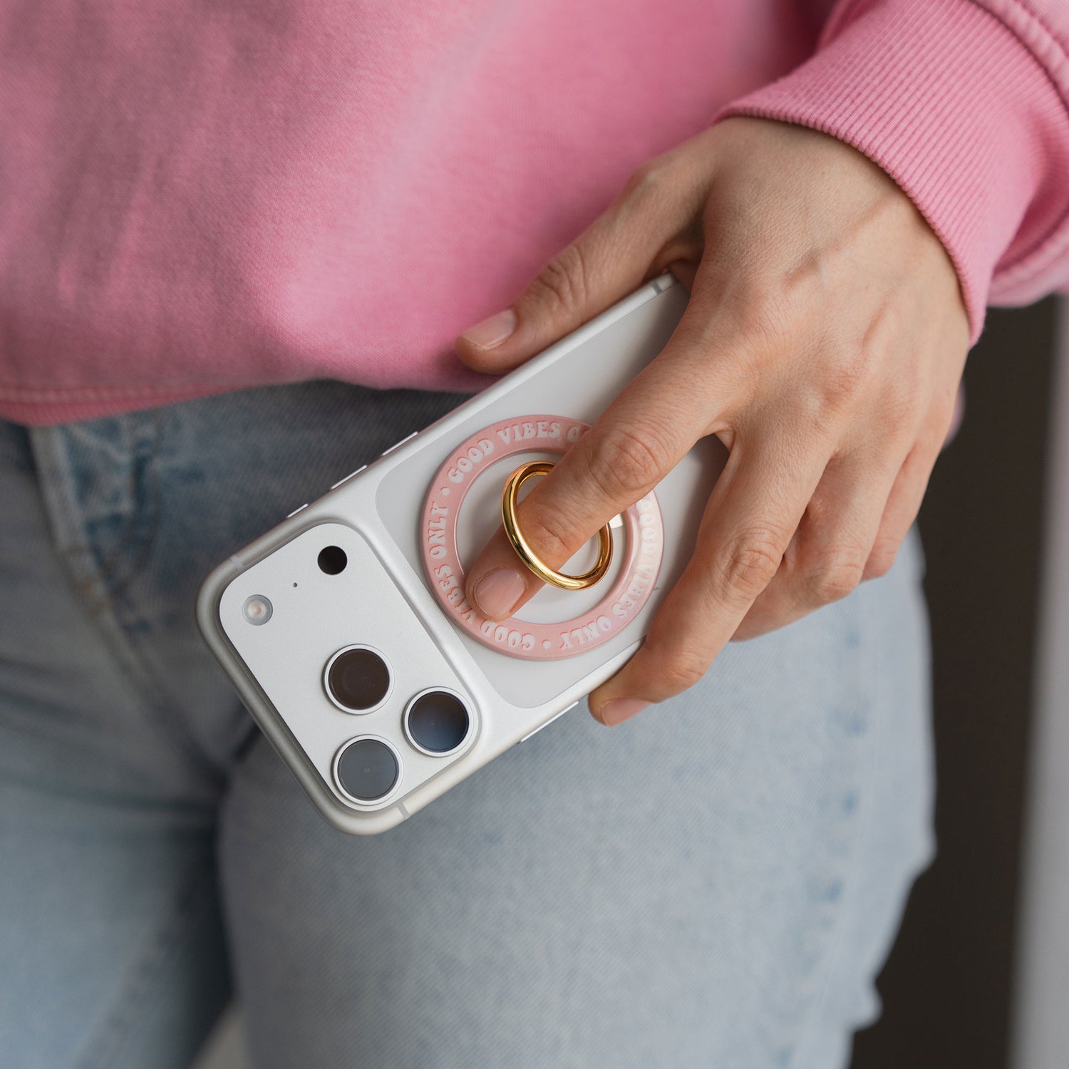 Eine Hand hält ein iPhone mit dem arktis Magnetischer Ringhalter für MagSafe - Good Vibes, getragen mit Jeans und einem rosa Sweatshirt - das ideale Zubehör für Ihren Stil.