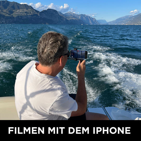 Ein Mann auf einem Boot filmt das Wasser mit seinem iPhone und fängt atemberaubende Berge und blauen Himmel ein - ideales Material für einen Online-Filmkurs der Wolffilms Academy von Rainer Wolf oder für die Erstellung professioneller Videos.