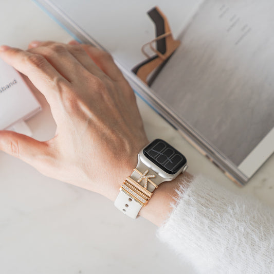 Eine Hand mit goldenen Ringen und Arktisband Apple Watch Charms „K Style“ ruht auf einem offenen Magazin und zeigt die Eleganz der Edelstahl- und Glasdetails von Arktisband.