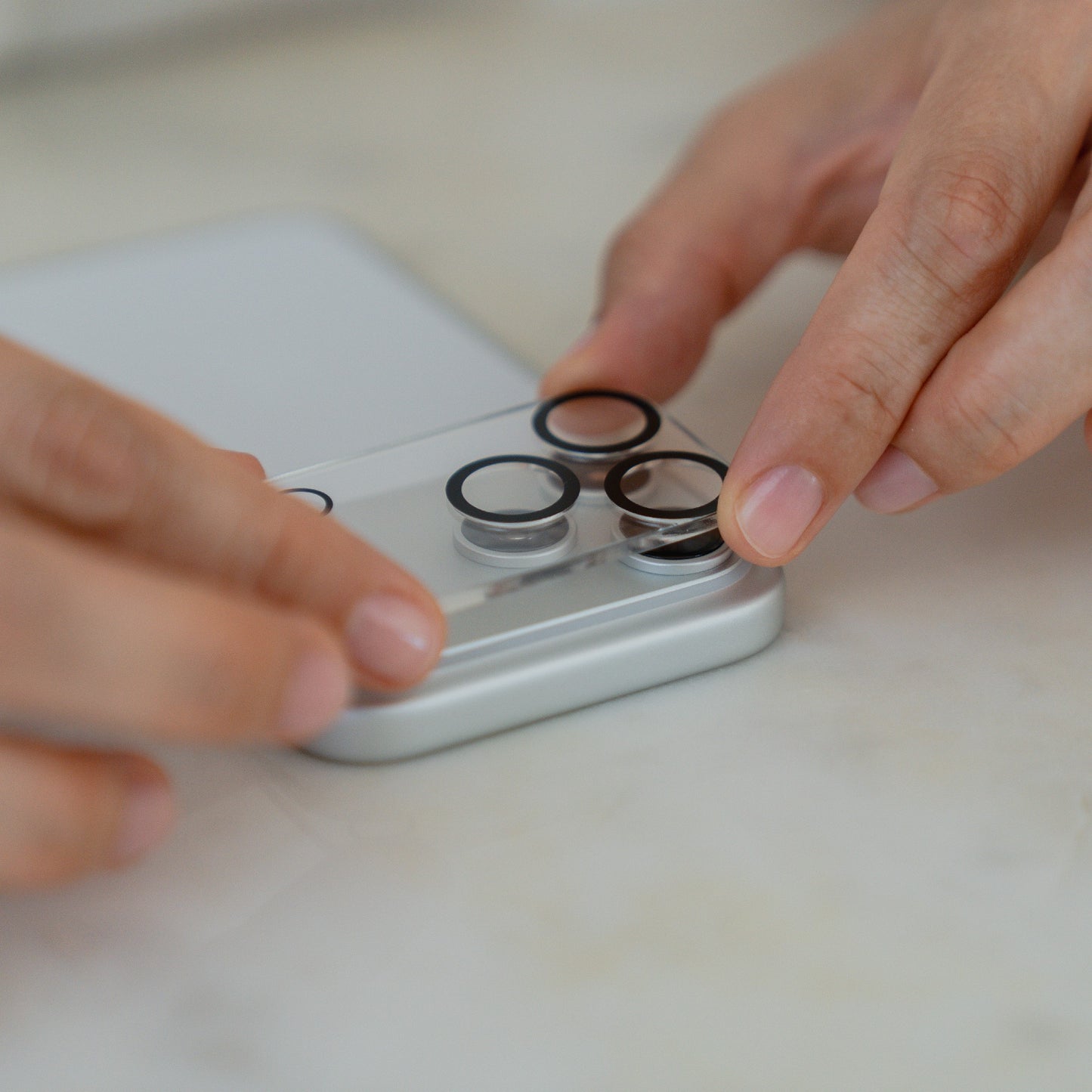 Nahaufnahme der Hände, die den arktis iPhone 17 Pro Max Kamera Protector von arktis auf die Kameralinsen eines iPhone 17 Pro Max auf einer weißen Fläche auftragen.