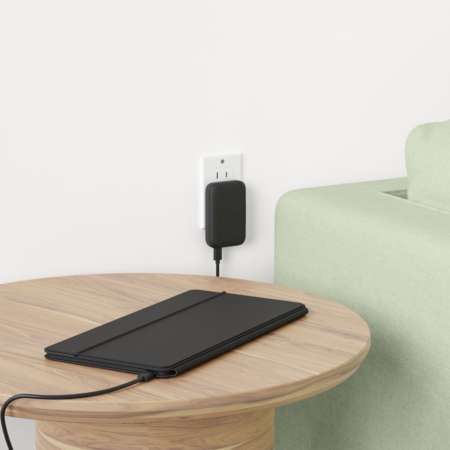 Ein Satechi OntheGo Bluetooth Keyboard mit Ständer von Satechi liegt auf einem runden Holztisch neben einer Steckdose hinter einem hellgrünen Sofa, neben einem Qi2 Magnetic Wireless Car Charger, der MagSafe kompatibel ist.