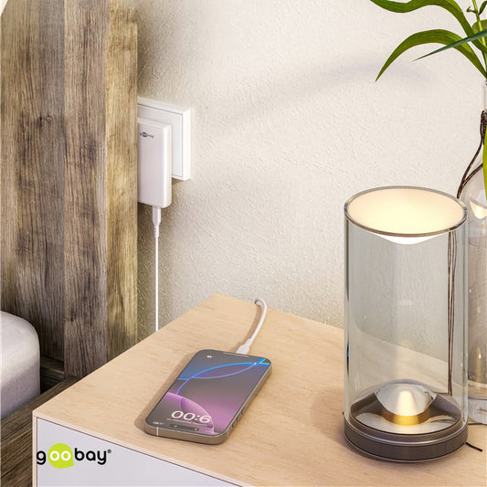 Ein Smartphone lädt auf einem Nachttisch neben einer modernen Lampe und einem eingesteckten Goobay USB-C Dual-Schnellladegerät 90° Flat 65 W.