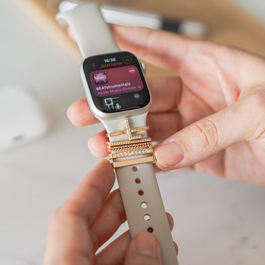Eine Hand passt ein helles Arktisband-Apple-Watch-Armband an, das mit Arktisband-Apple-Watch-Charms „L Style“ verziert ist. Auf dem Bildschirm der Smartwatch wird eine Musik-App mit einer BEATstrumentals-Playlist angezeigt.