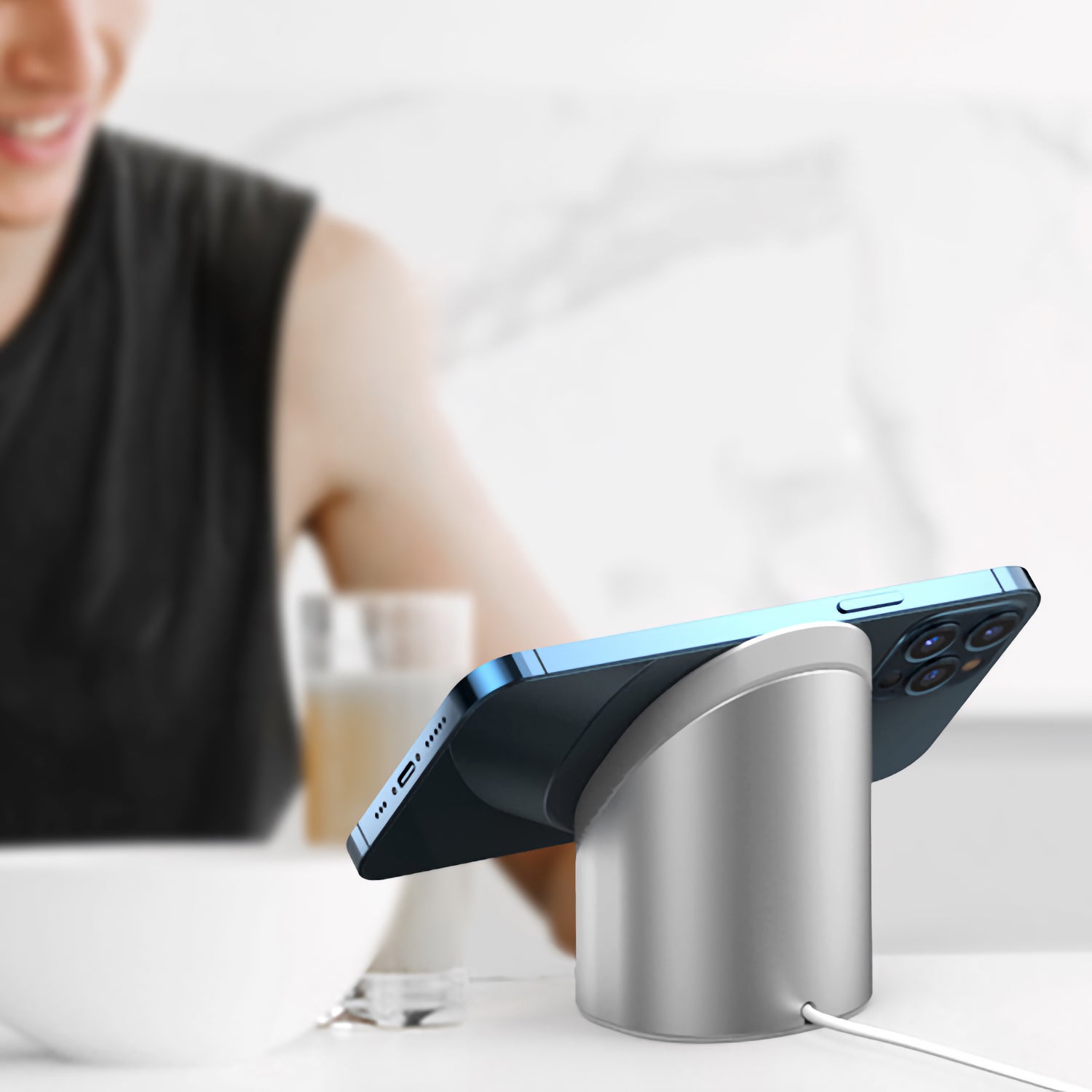 Ein blaues Smartphone ruht auf einem arktis MagSafe NightStand, mit einer lächelnden Person und einem Getränk im Hintergrund.