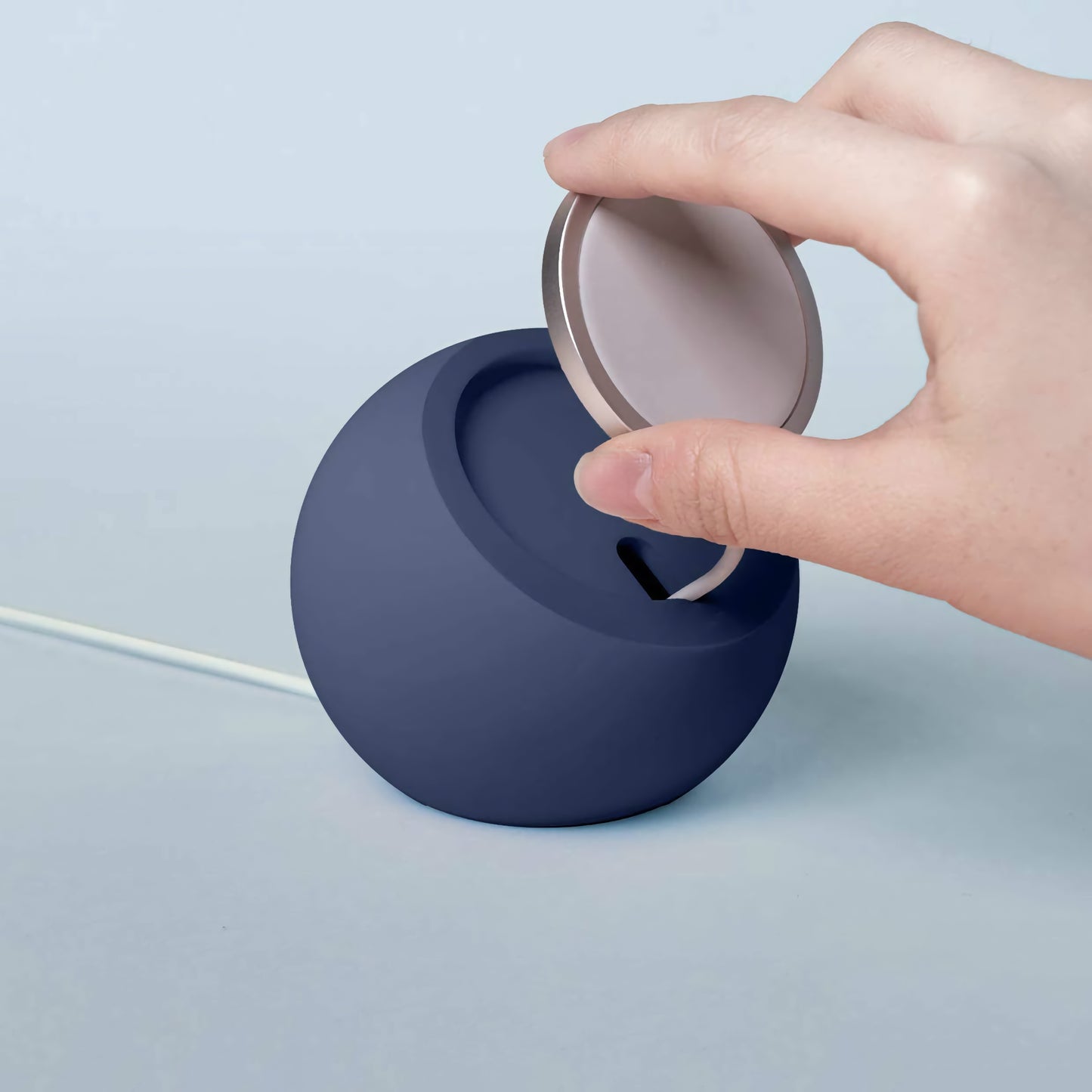 Eine Hand hebt den Deckel eines runden, dunkelblauen arktis MagSafe Orbit Stands mit angeschlossenem Kabel, perfekt für kabelloses Laden.