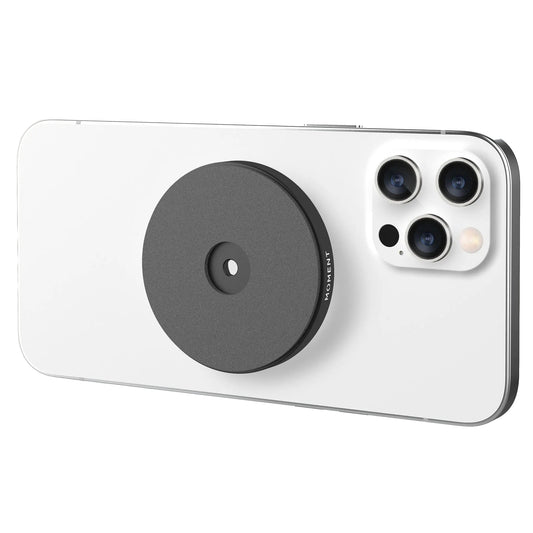Ein weißes Smartphone mit einer MOMENT Moment Wall Mount für MagSafe (schwarz, rund), die in der Nähe des dreifachen Kameramoduls auf der Rückseite angebracht ist.