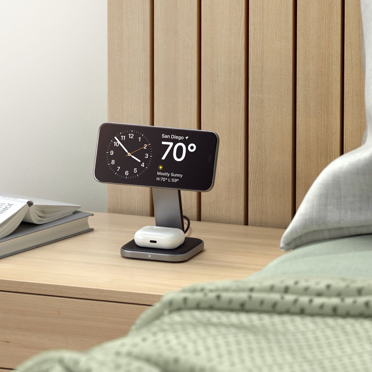 Der Satechi 2-in-1 Foldable Qi2 Wireless Charging Stand hält ein Telefon, das die Uhrzeit und das Wetter anzeigt, auf dem Nachttisch, zusammen mit einem Buch und einer AirPods-Ladestation.