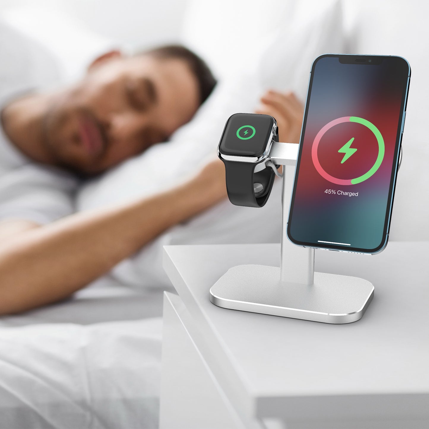 Ein iPhone und eine Apple Watch laden auf dem arktis MagSafe AluStand für iPhone und Apple Watch von arktis, neben einem schlafenden Mann im Bett.