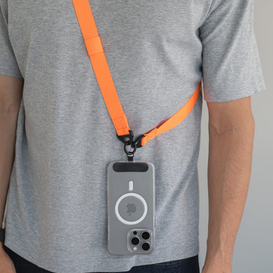 Eine Person in einem hellgrauen T-Shirt trägt ein iPhone in einer durchsichtigen Hülle, die an einem orangefarbenen Arktis-Smartphone-Gurtband mit 1-Klick-Verschluss befestigt ist. Es wird quer über den Körper getragen und an der Hülle befestigt, sodass das Telefon vorne hängt.