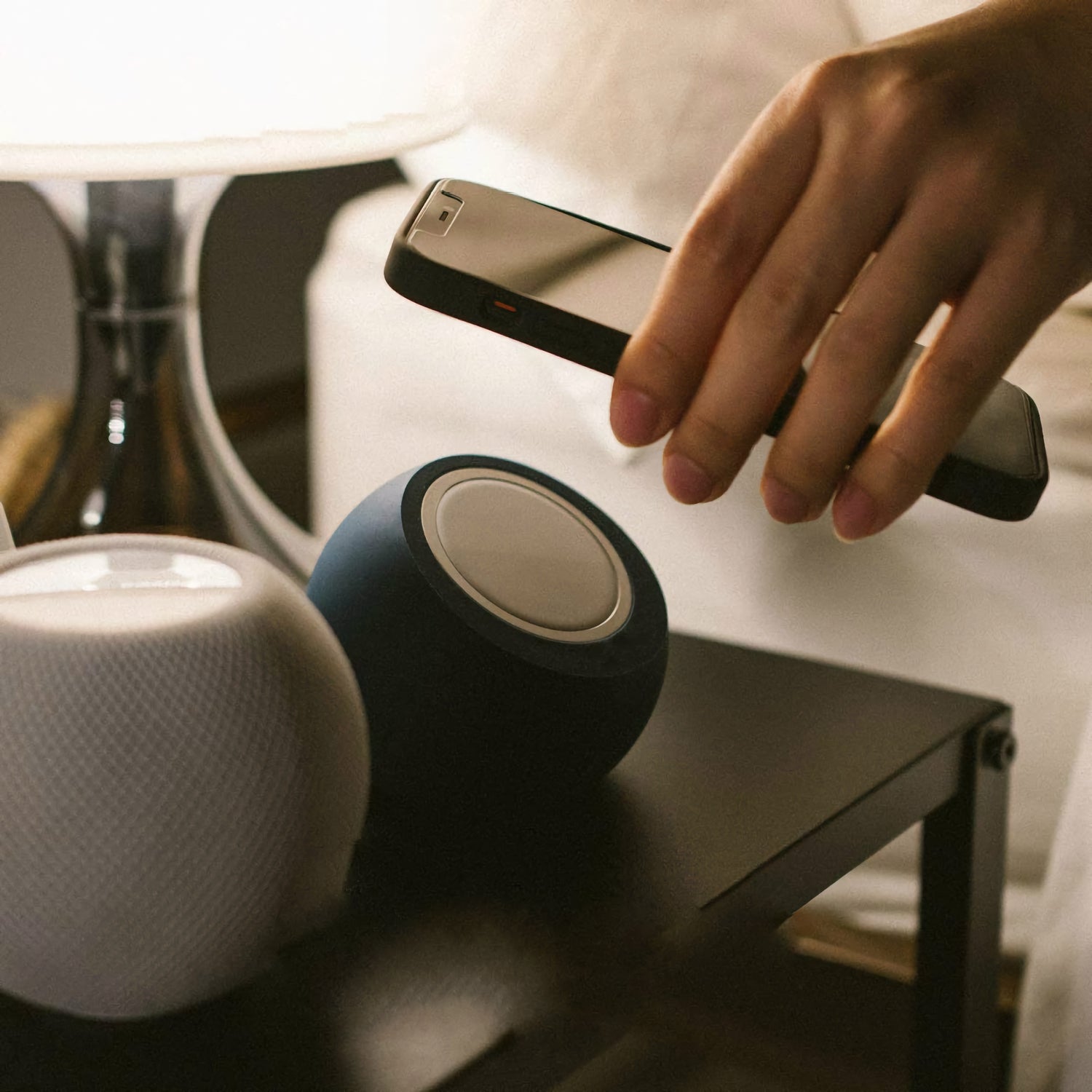 Eine Hand hält ein Smartphone neben zwei Smart Speakern und dem arktis MagSafe Orbit Stand von arktis auf einem Nachttisch neben einer Lampe.