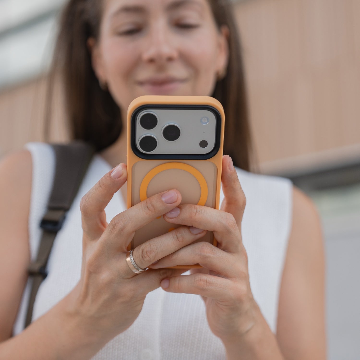 Eine Frau lächelt auf ihr Smartphone, das sie mit beiden Händen in einer orangefarbenen, matten Arktis arktis iPhone 17 Pro Solid Case mit MagSafe hält - ihre elegante Hülle von Arktis ermöglicht müheloses Laden mit MagSafe.