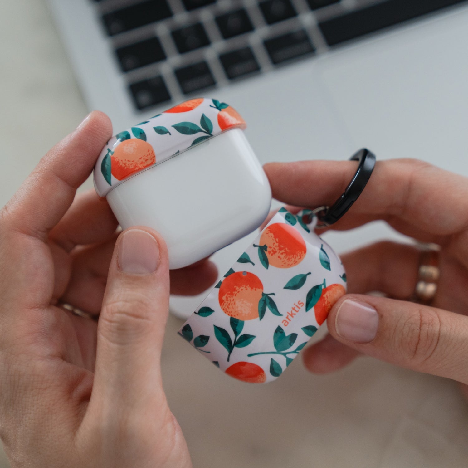 Hände, die eine Arktis AirPods 4-Hülle – das Orange Tree-Case – mit einem Muster aus Orangen und grünen Blättern halten.