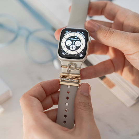 Eine Person passt eine Smartwatch mit beigem Arktisband-Armband an, das mit gold- und diamantbesetzten Arktisband Apple Watch Charms „P Style“ verziert ist. Das digitale Zifferblatt ist zu sehen, im Hintergrund ein unscharfes Buch und eine Brille.