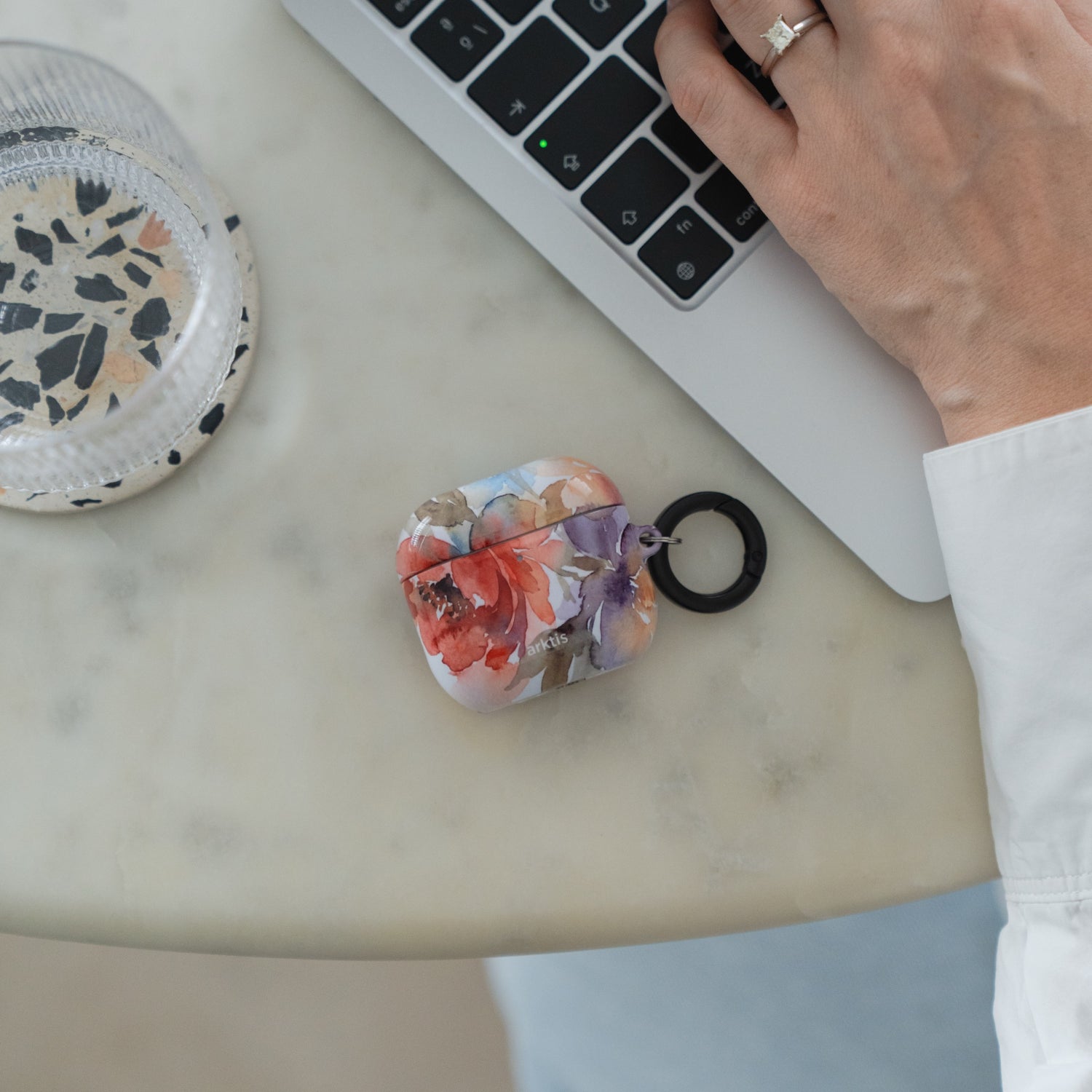Arktis AirPods 5-Hülle – Floral mit schwarzem Befestigungsring, die auf einem Tisch neben einem Laptop liegt.