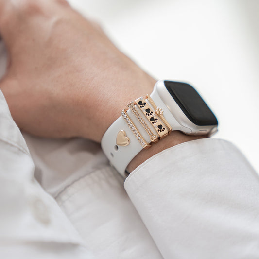 Eine Person trägt die arktisband Apple Watch Charms „Pet Love“ in Gold – mit Herz-, Pfotenabdruck- und Kronendesign – als einzigartige Charms auf ihrer Apple Watch über einem weißen Langarmshirt.