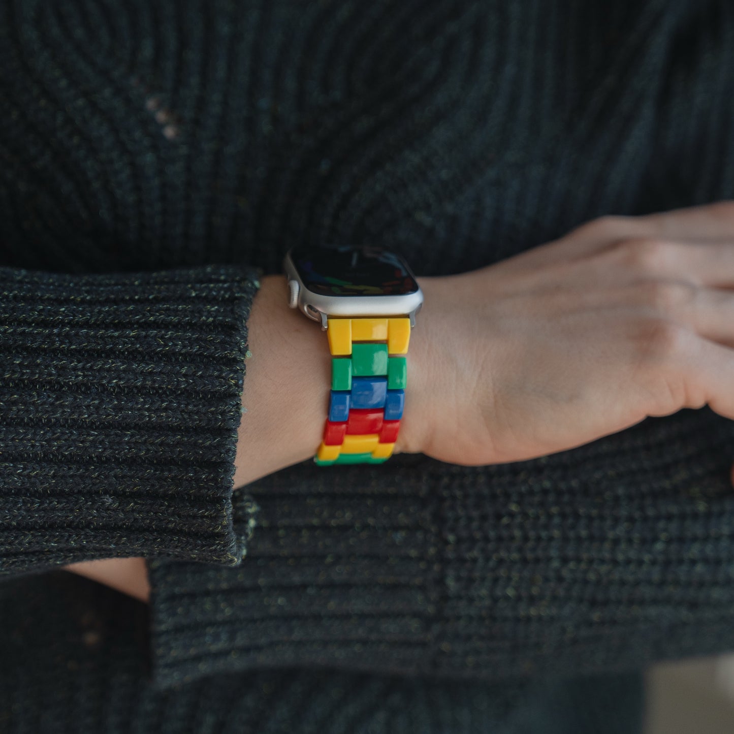 Jemand trägt eine Smartwatch mit dem arktisband Apple Watch "Pixel Band", einem farbenfrohen Armband im Retrostil mit Blockdesign, das über einem dunklen Strickpullover getragen wird.