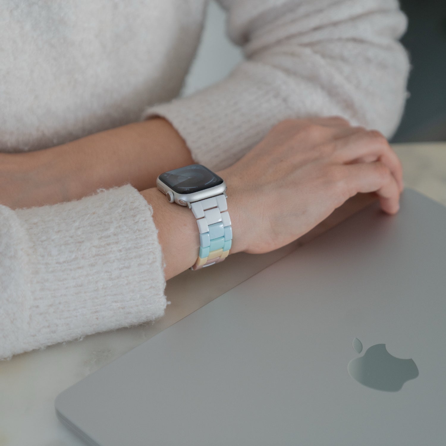 Jemand, der die Apple Watch "Pixel Band" von arktisband mit einem pastellfarbenen Band trägt, stützt seine Hand auf einen geschlossenen silbernen Laptop.