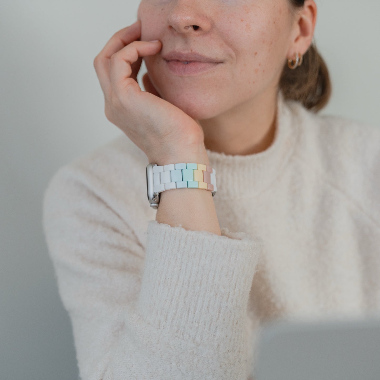 Eine Frau in einem cremefarbenen Pullover stützt ihr Kinn auf ihre Hand und trägt die Apple Watch "Pixel Band" von arktisband in Retro-Pastellfarben.