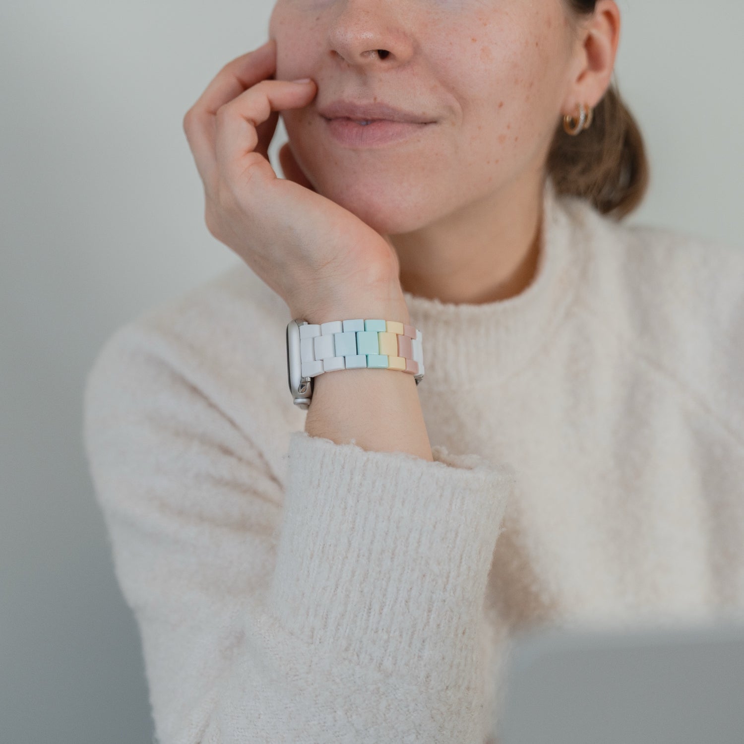 Eine Frau in einem cremefarbenen Pullover stützt ihr Kinn auf ihre Hand und trägt die Apple Watch "Pixel Band" von arktisband in Retro-Pastellfarben.