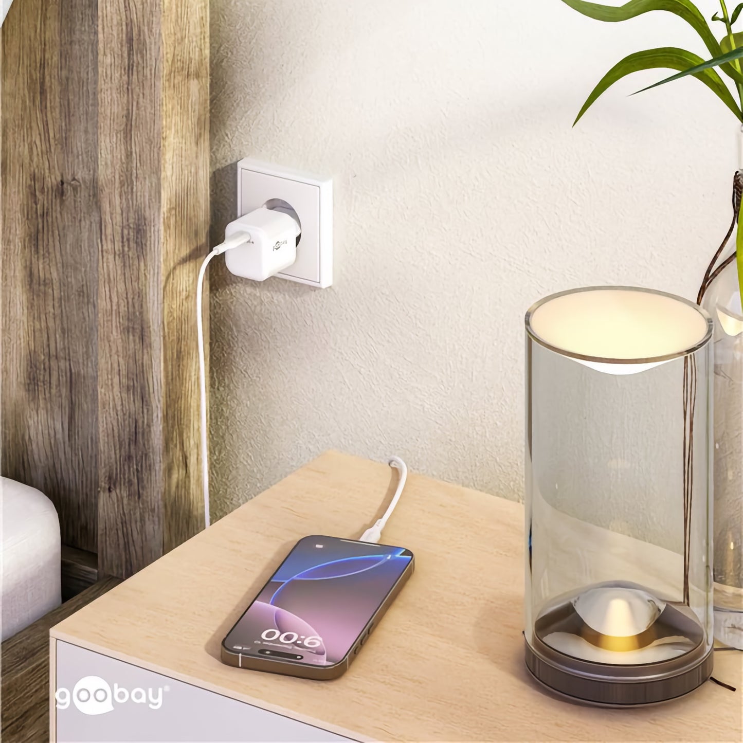 Ein Smartphone wird auf einem Nachttisch neben einer Lampe aufgeladen, eingesteckt in die Wand mit einem Goobay USB-C PD GaN Schnellladegerät Nano 30 W.