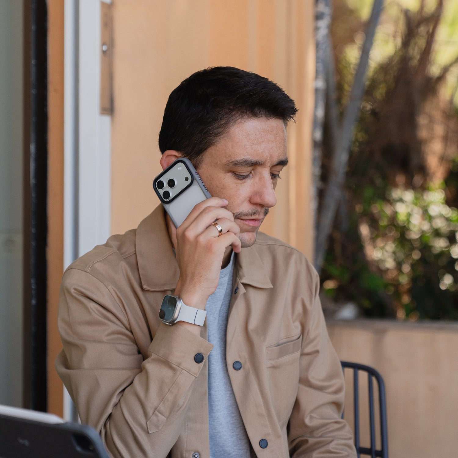 Ein Mann mit hellbrauner Jacke sitzt im Freien und spricht mit seinem Smartphone, geschützt durch das Arktis iPhone 17 Pro Solid Case, eine matte, stoßabsorbierende Hülle von Arktis.