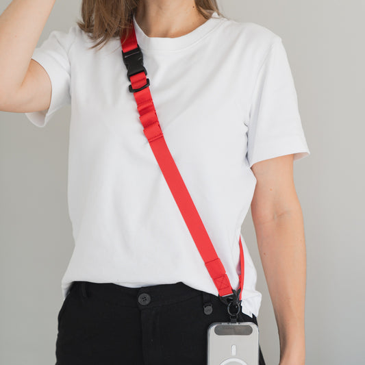 Eine Person in einem weißen T-Shirt und einer schwarzen Hose trägt ein rotes arktis Smartphone-Gurtband-Lanyard mit 1-Klick-Verschluss quer über dem Oberkörper, das an einer seitlich an ihr hängenden Smartphone-Hülle befestigt ist.