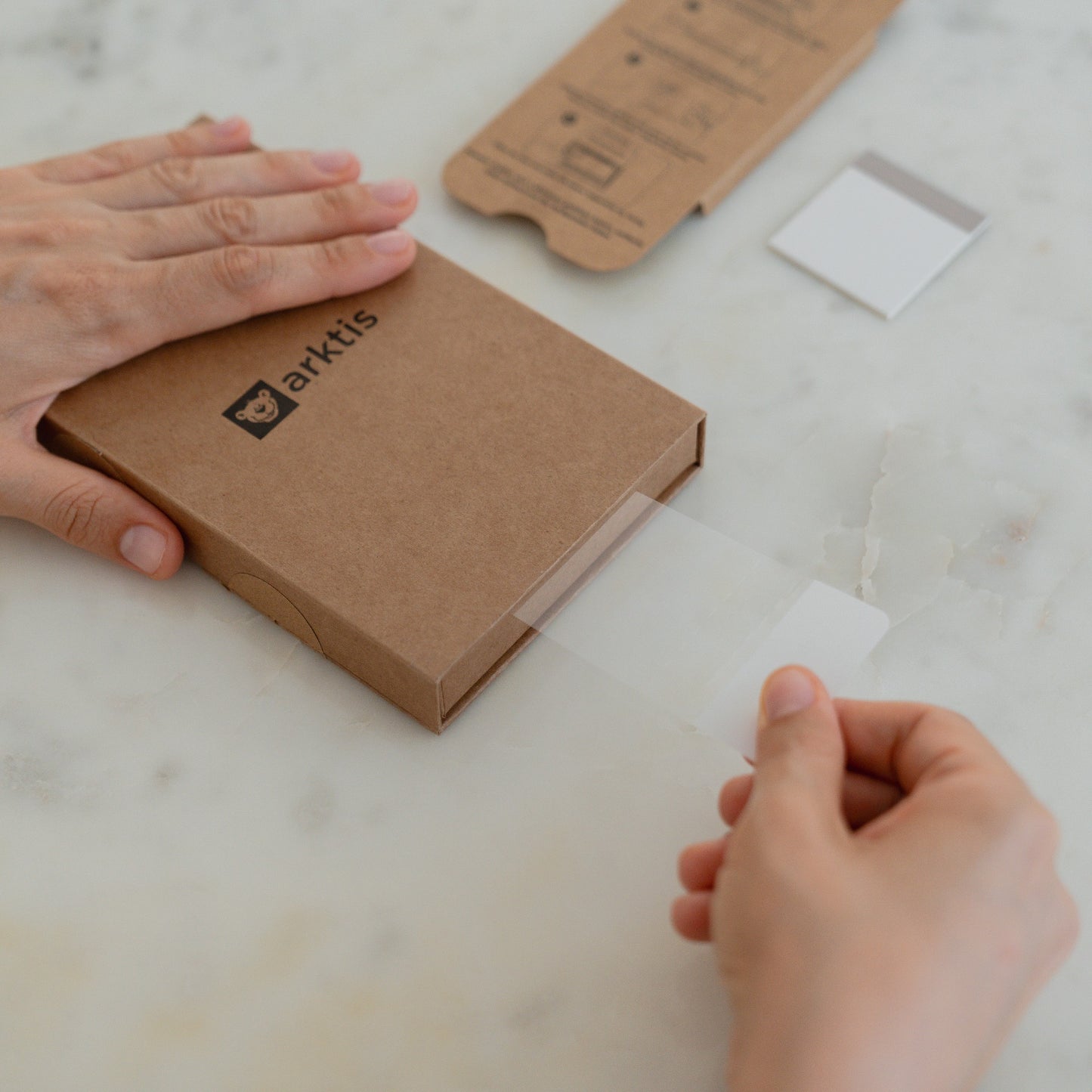 Hände, die ein transparentes arktis 2er Set iPhone 17 Pro Max REVOLUTION Glas-Displayschutz aus einer braunen Schachtel auf einer weißen Marmorfläche entnehmen, wahrscheinlich für das neue iPhone 17 Pro Max.