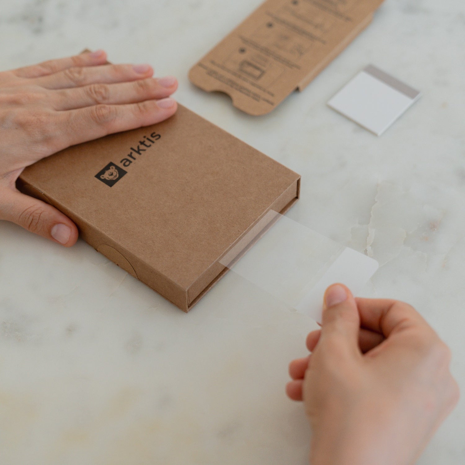 Hände, die ein transparentes arktis 2er Set iPhone 17 Pro Max REVOLUTION Glas-Displayschutz aus einer braunen Schachtel auf einer weißen Marmorfläche entnehmen, wahrscheinlich für das neue iPhone 17 Pro Max.