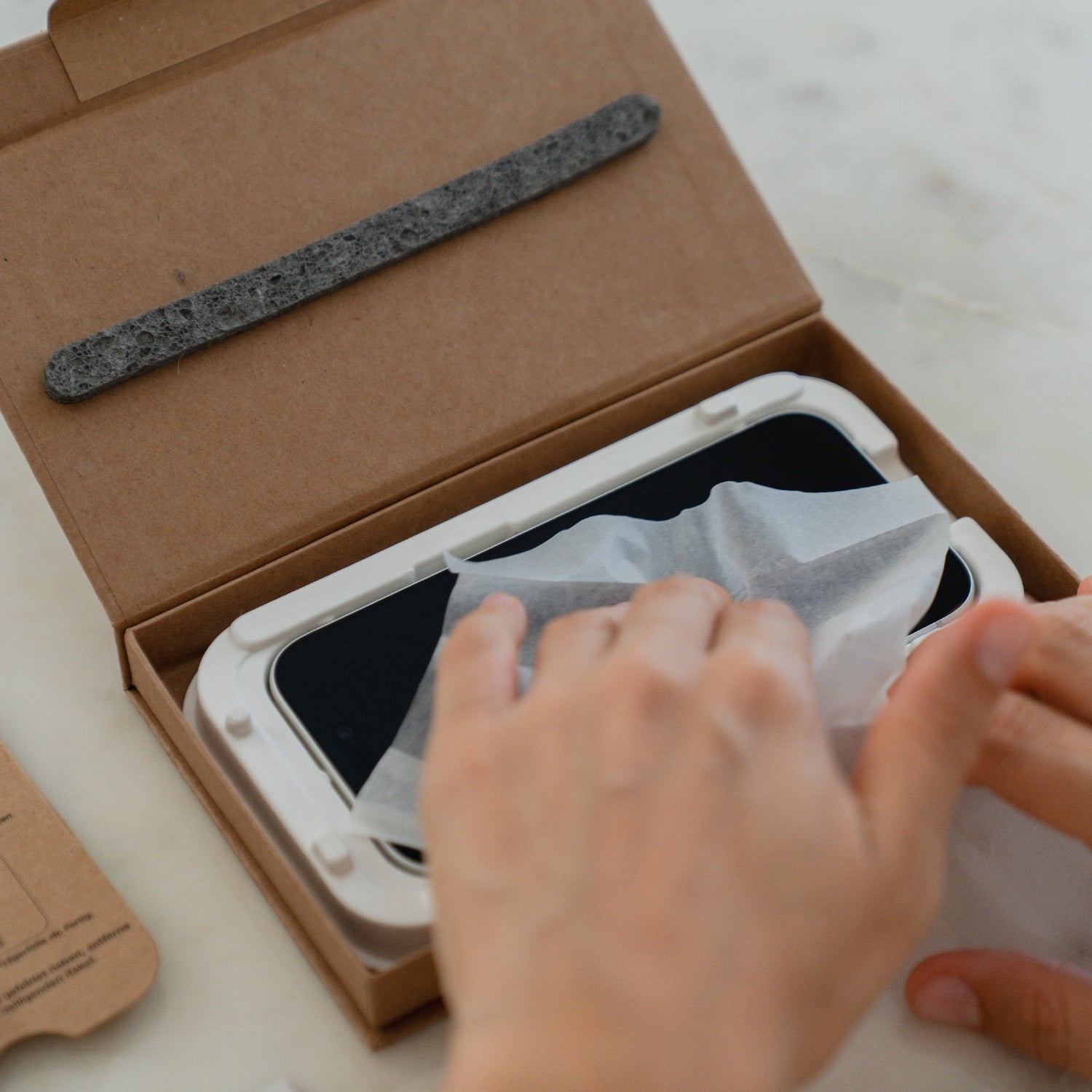 Hände, die ein Smartphone mit einem Taschentuch abwischen; das Telefon, geschützt durch einen arktis 2er Set iPhone 17 Pro Max REVOLUTION Glas-Displayschutz, ruht in einer EZ.INSTALL Box mit einem schwarzen Streifen darüber.
