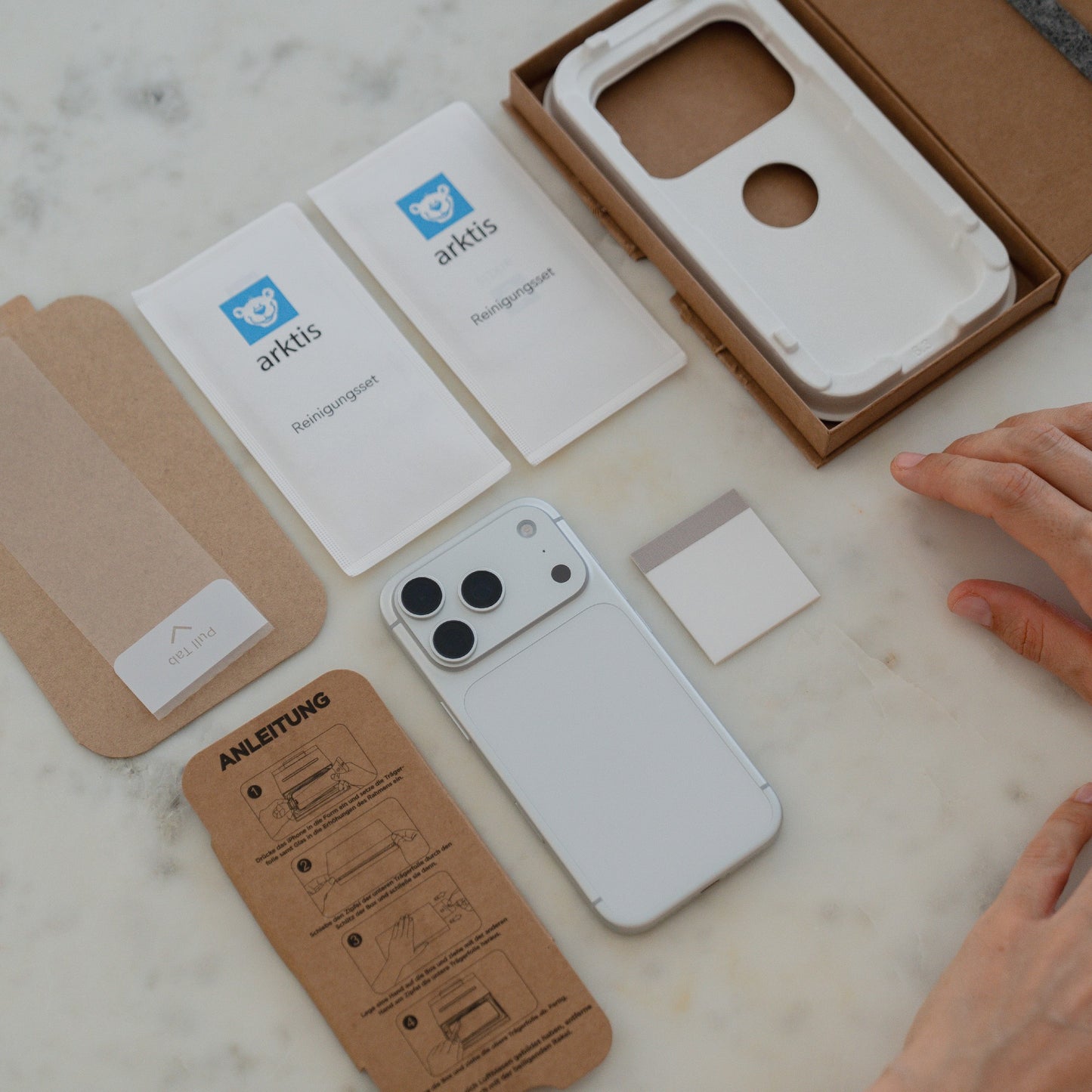 Das arktis 2er Set iPhone 17 Pro Max REVOLUTION Glas-Displayschutz, Reinigungstücher und Anleitung sind auf einer Marmorfläche angeordnet, mit den Händen wird die Montage des Glas-Displayschutzes vorbereitet.