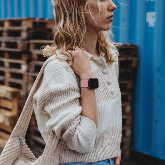 Eine Person mit welligem blondem Haar, einem creme-weiß gestreiften Pullover und Bluejeans, trägt eine beige Cordtasche und trägt das arktisband Apple Watch Magnetarmband „Neo“, während sie neben gestapelten Paletten und einem blauen Schiffscontainer steht.