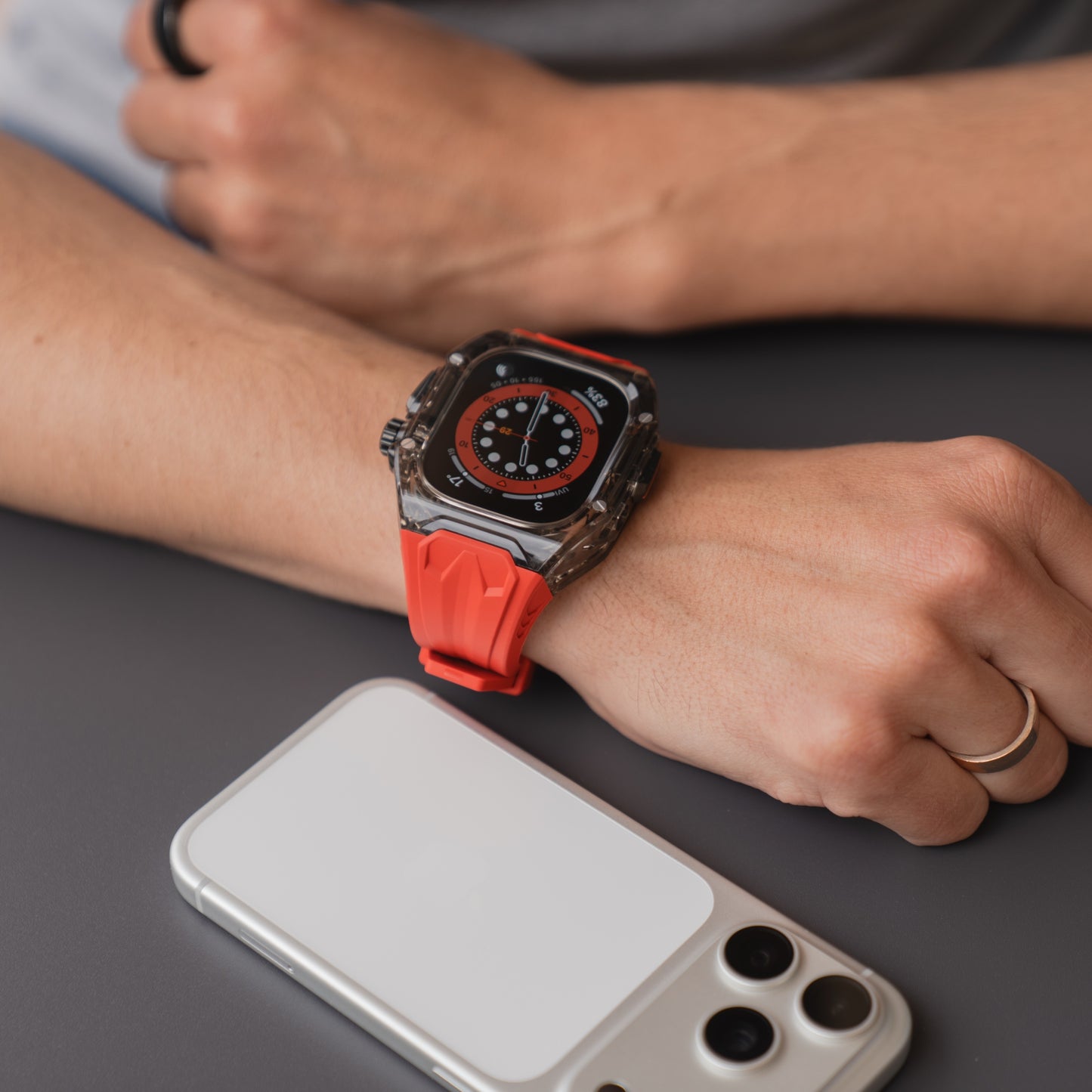 Eine Person, die ein arktisband Apple Watch Armband "Armor Fusion" mit rotem Band trägt, sitzt neben einem Smartphone auf einer grauen Fläche.
