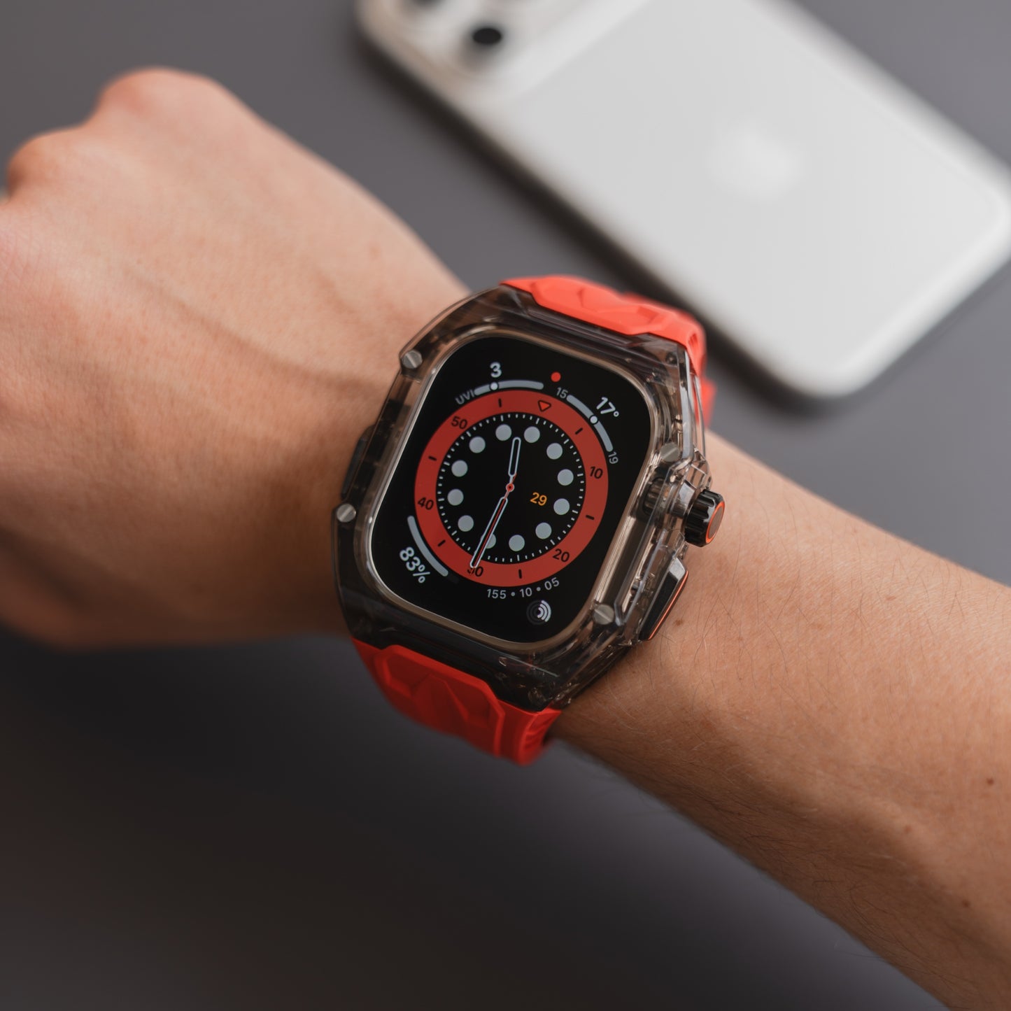 Eine Person, die das arktisband Apple Watch Armband "Armor Fusion" mit einem roten Silikonband trägt, steht neben einem Smartphone in einem transparenten Hardcase auf einer grauen Fläche.