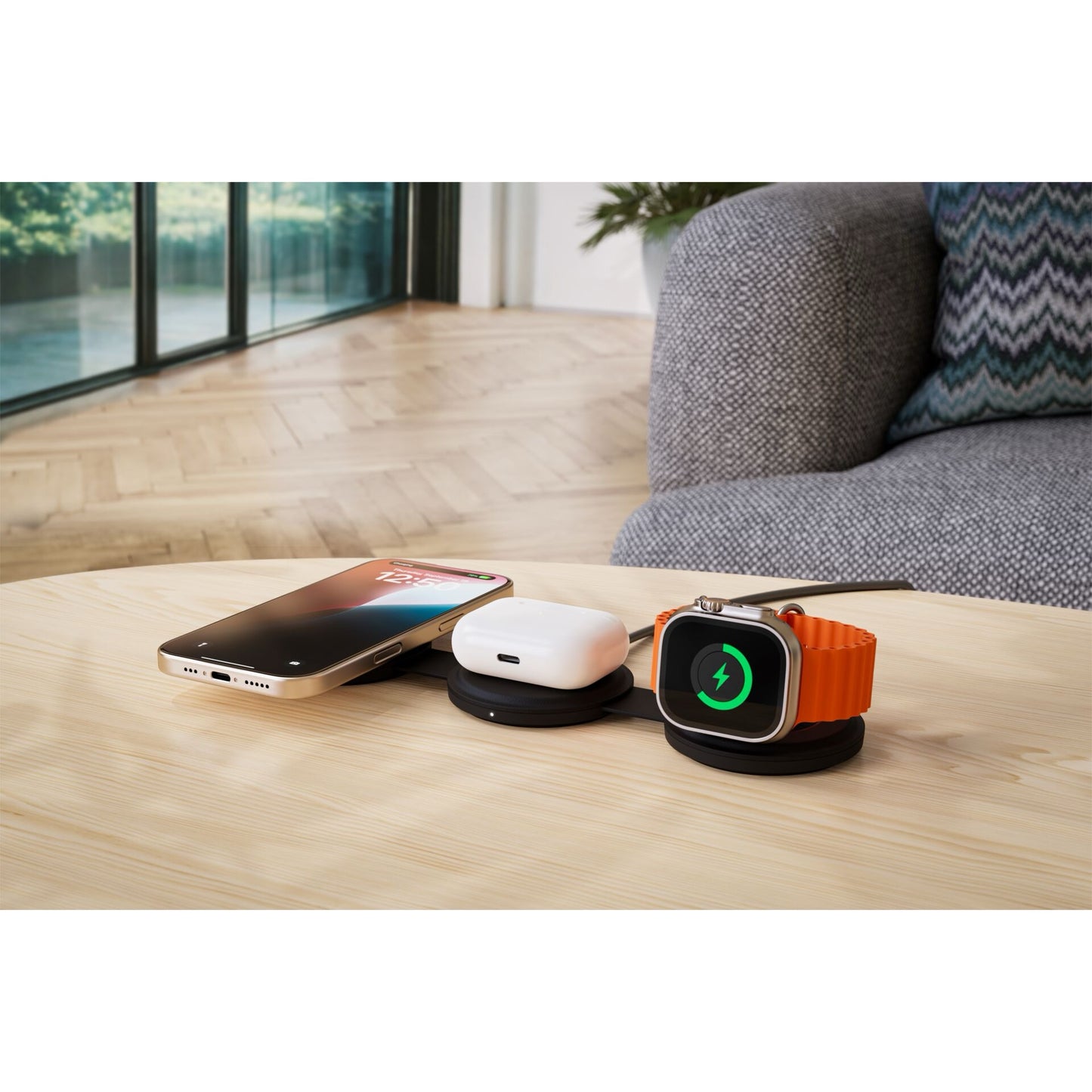 Ein Satechi OntheGo 3-in-1 Wireless Charger versorgt ein Smartphone, Ohrstöpsel und eine Smartwatch mit Qi2-Technologie auf einem Holztisch neben einer grauen Couch in einem modernen Wohnzimmer.